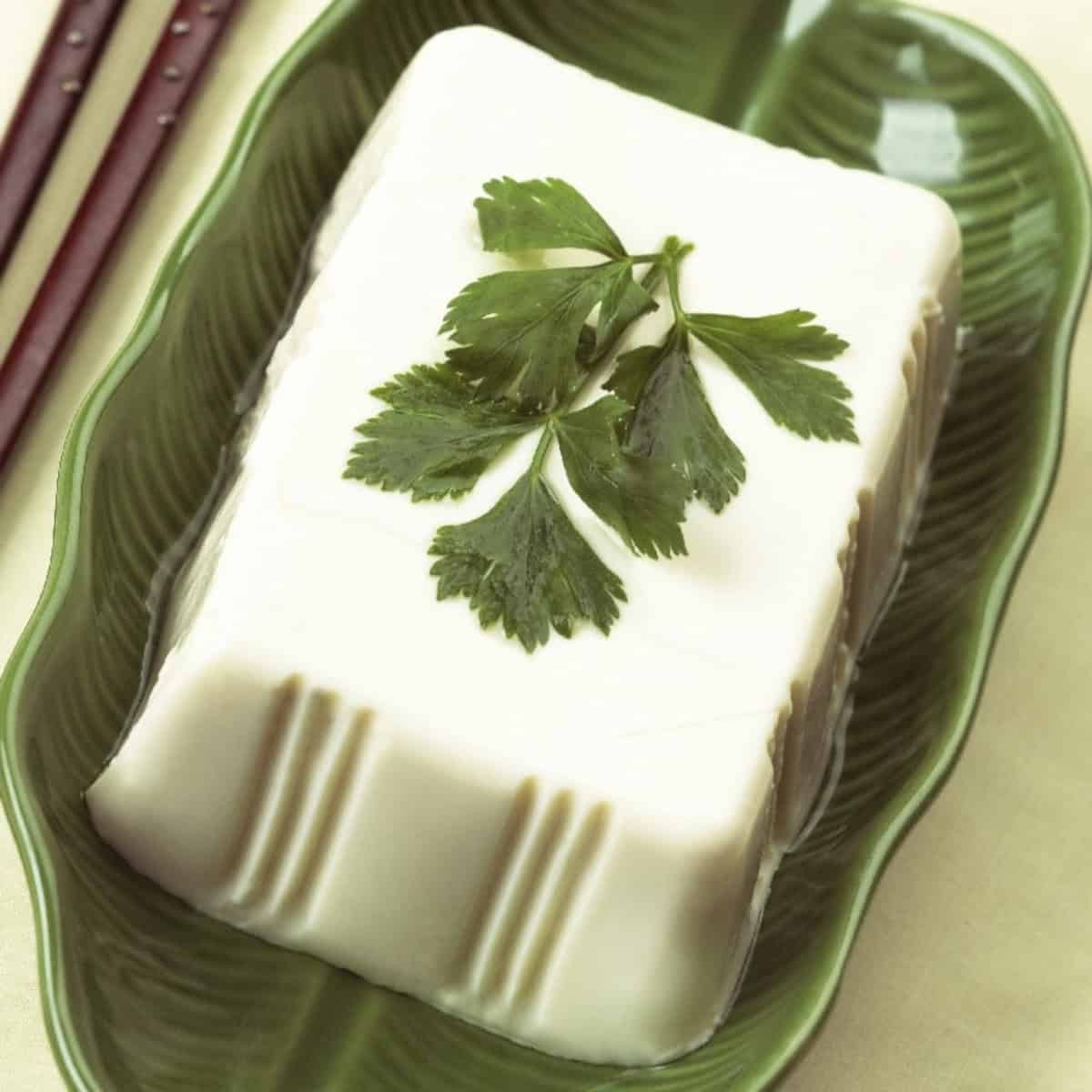 Silken tofu block on a leaf-shaped plate with chopsticks, a popular plant-based egg substitute.