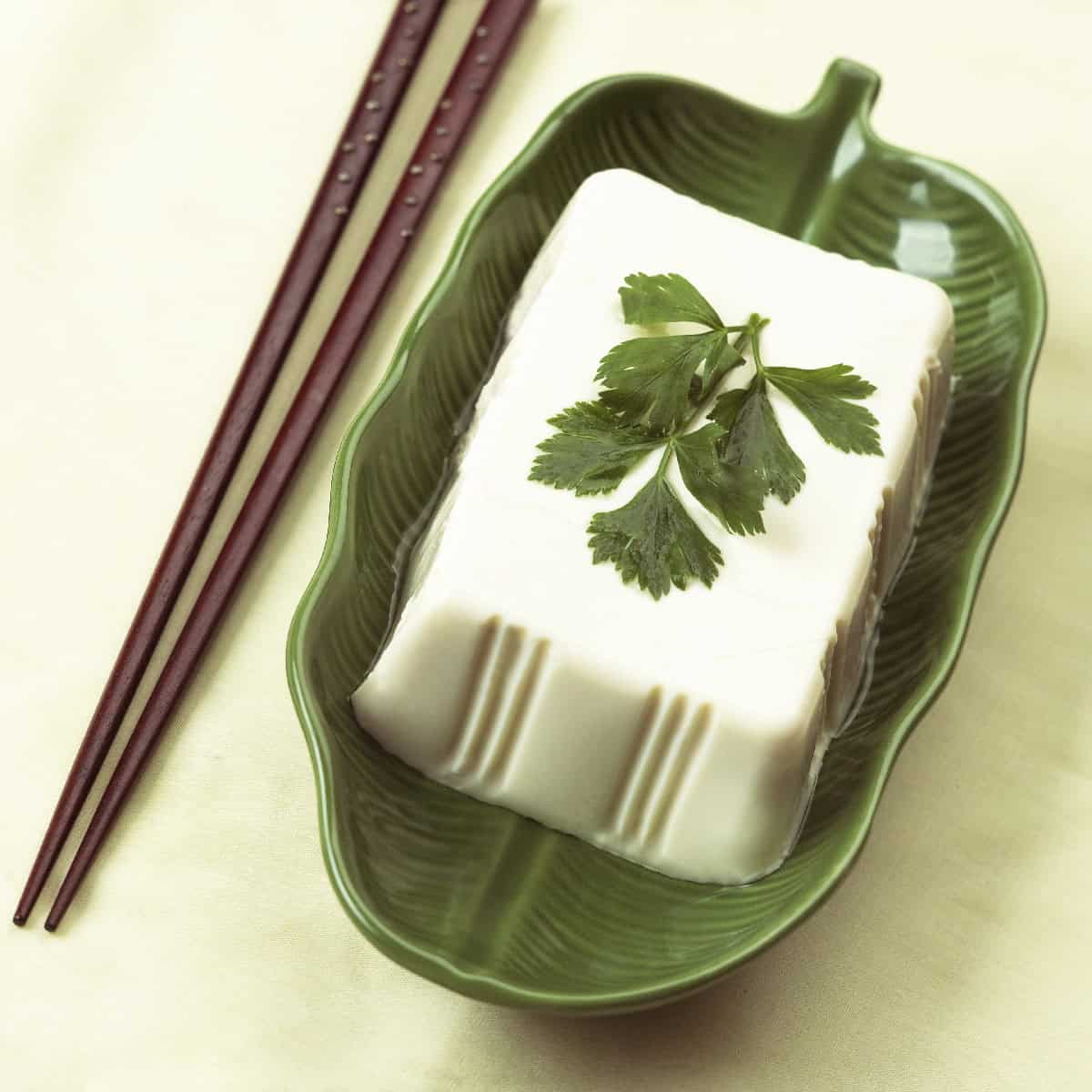 Silken tofu block on a green leaf-shaped plate with chopsticks, a popular plant-based egg substitute.