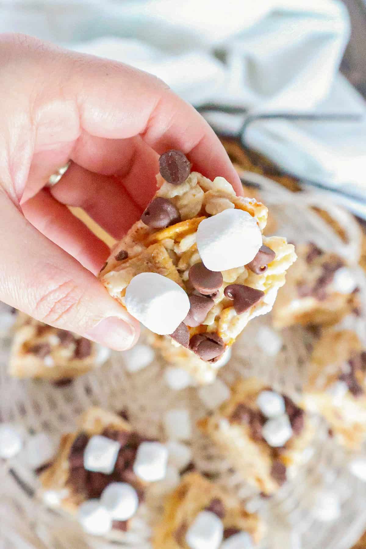Hand holding a homemade S'mores Rice Krispie treat topped with chocolate chips and mini marshmallows.