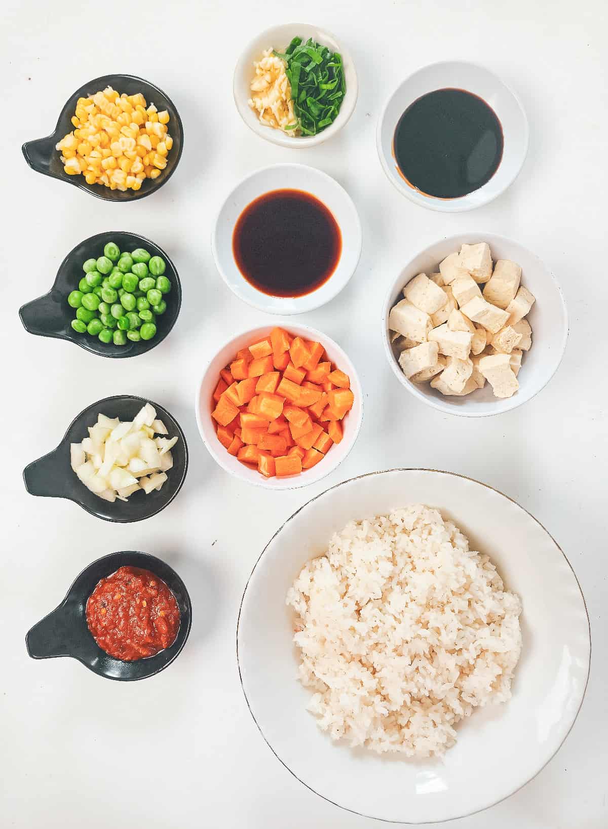 Ingredients for vegan nasi goreng, including cooked rice, tofu, carrots, peas, corn, onion, garlic, green onions, soy sauce, and sambal.