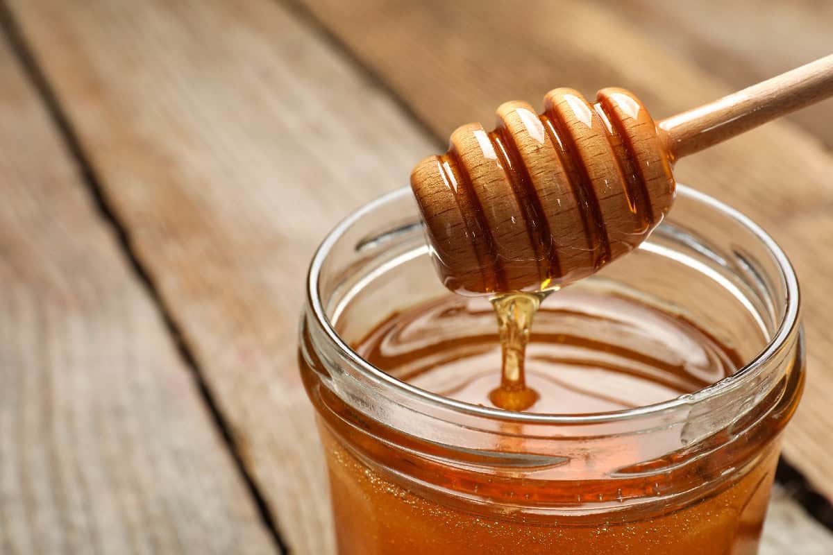 Honey dripping from a wooden dipper into a glass jar on a rustic wooden table.