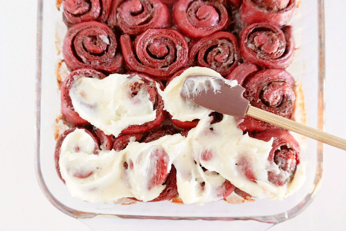 Baked red velvet cinnamon rolls being topped with cream cheese frosting.