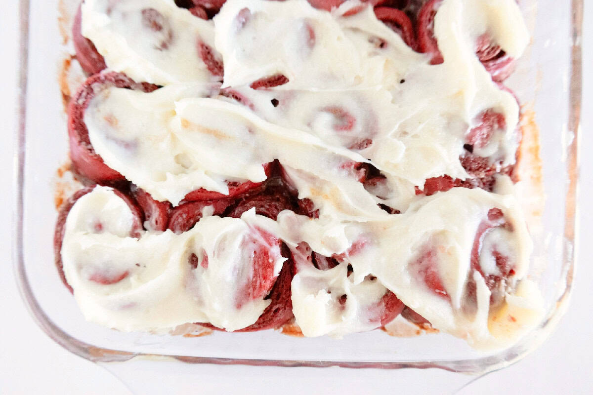 Baked red velvet cinnamon rolls being topped with cream cheese frosting.