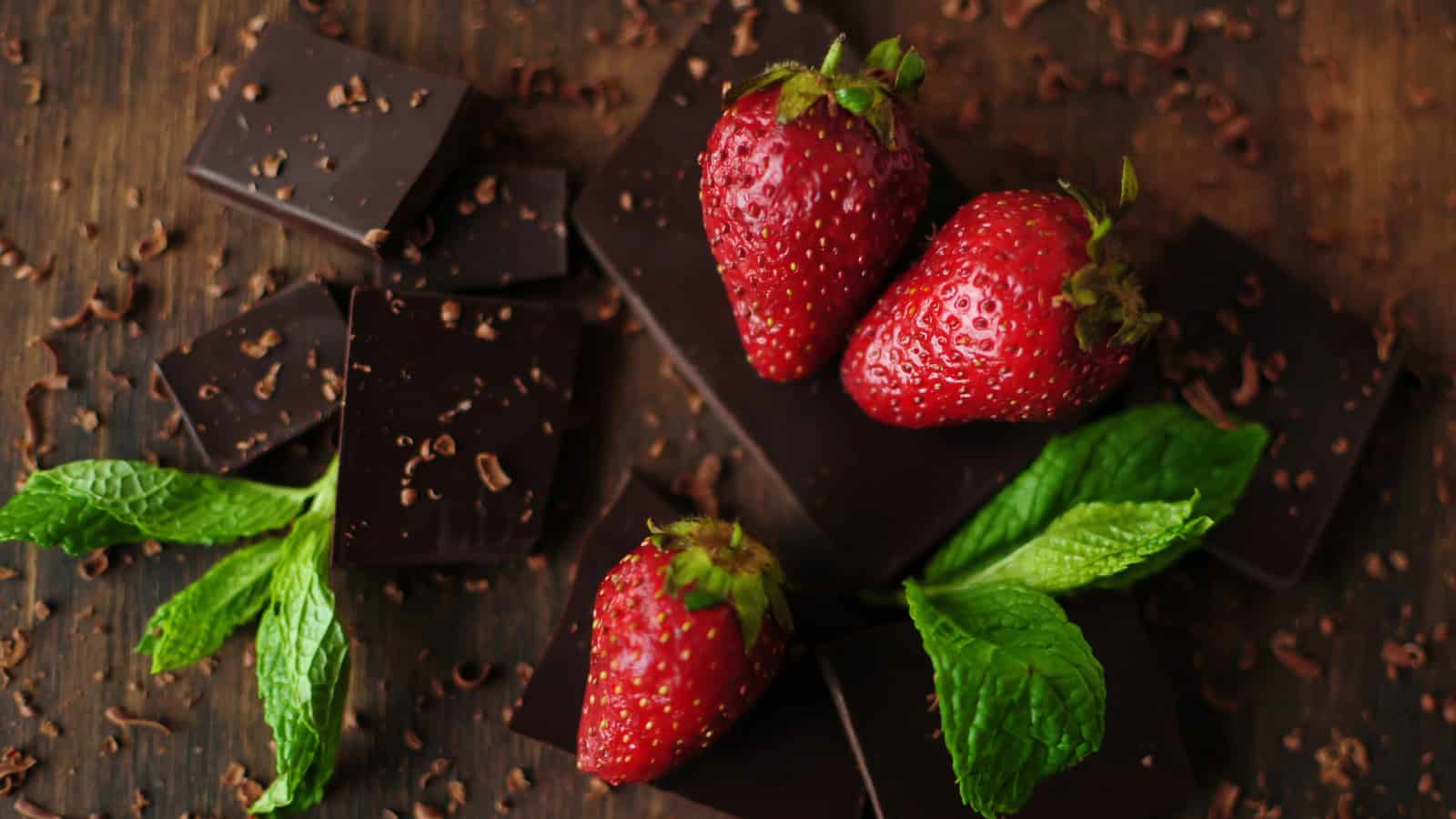 Fresh strawberries and mint leaves arranged with dark chocolate pieces on a wooden surface.