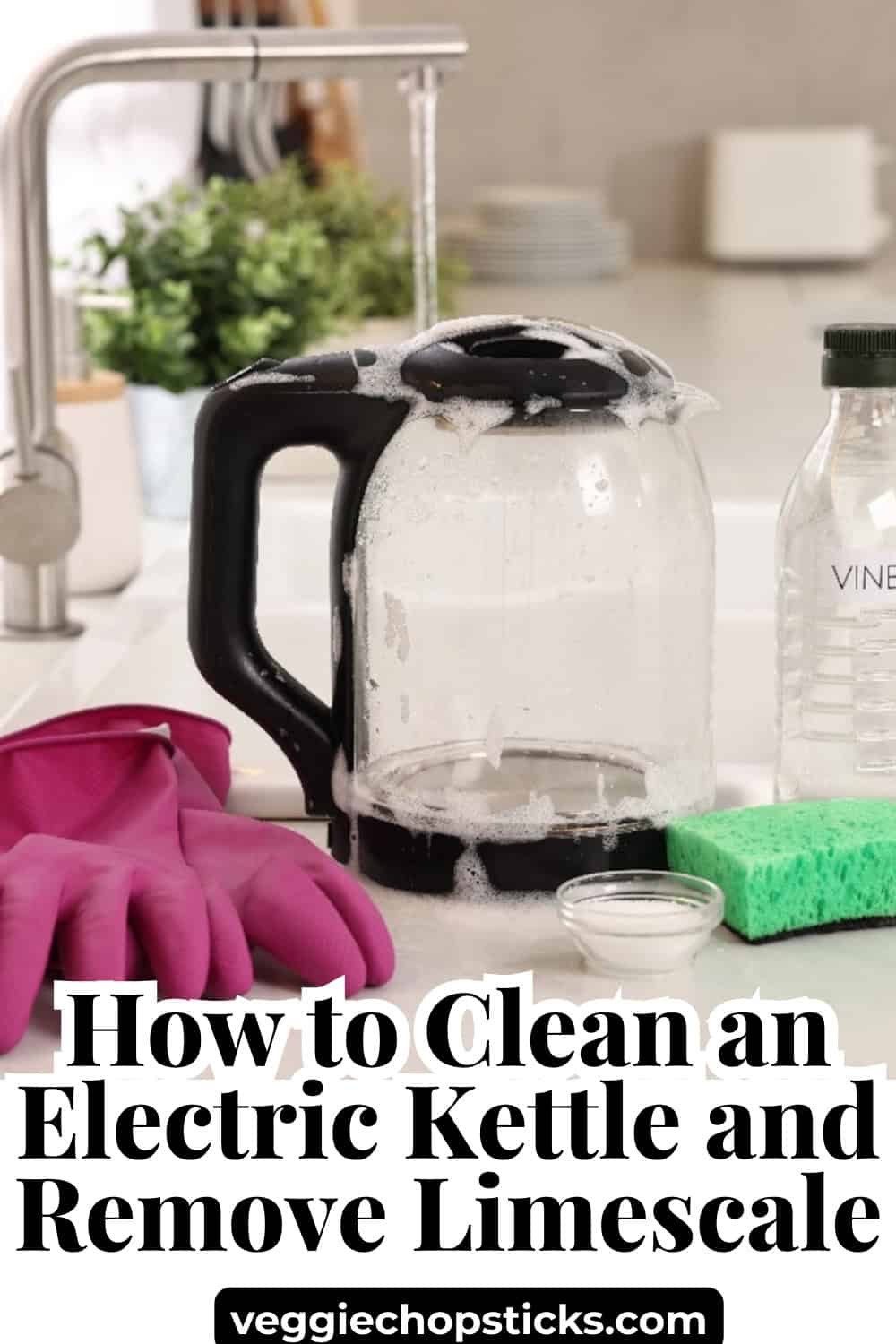 A glass electric kettle covered in soap suds next to cleaning gloves, vinegar, and a sponge.