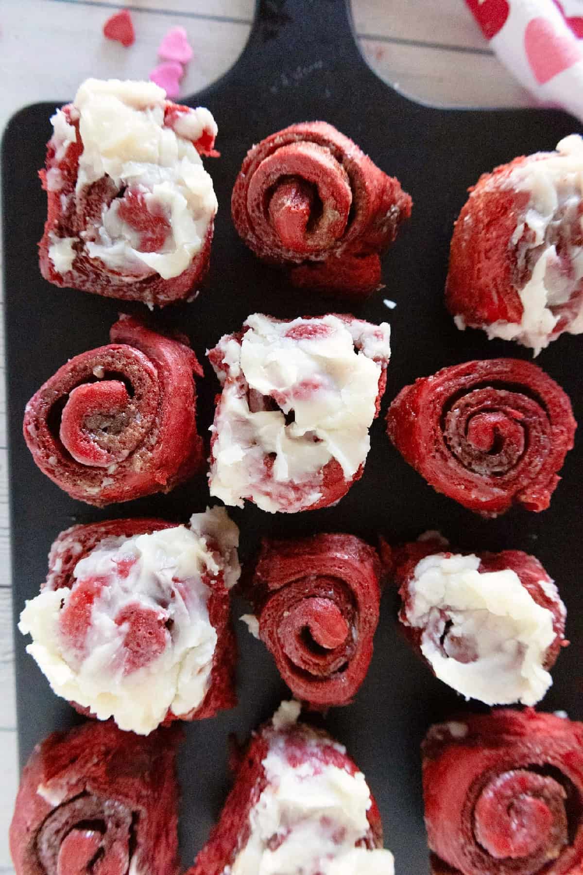 Red velvet cinnamon rolls with cream cheese frosting arranged on a black serving board.