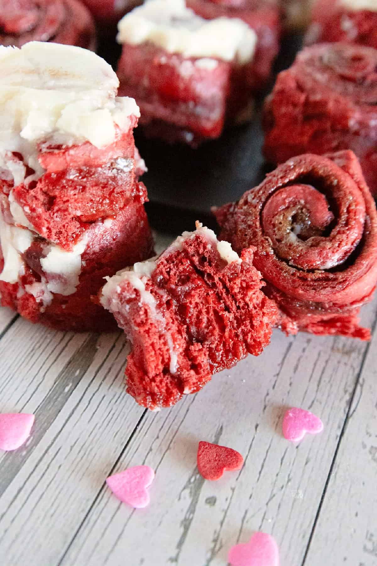 Red velvet cinnamon rolls with cream cheese frosting surrounded by pink and red heart sprinkles.
