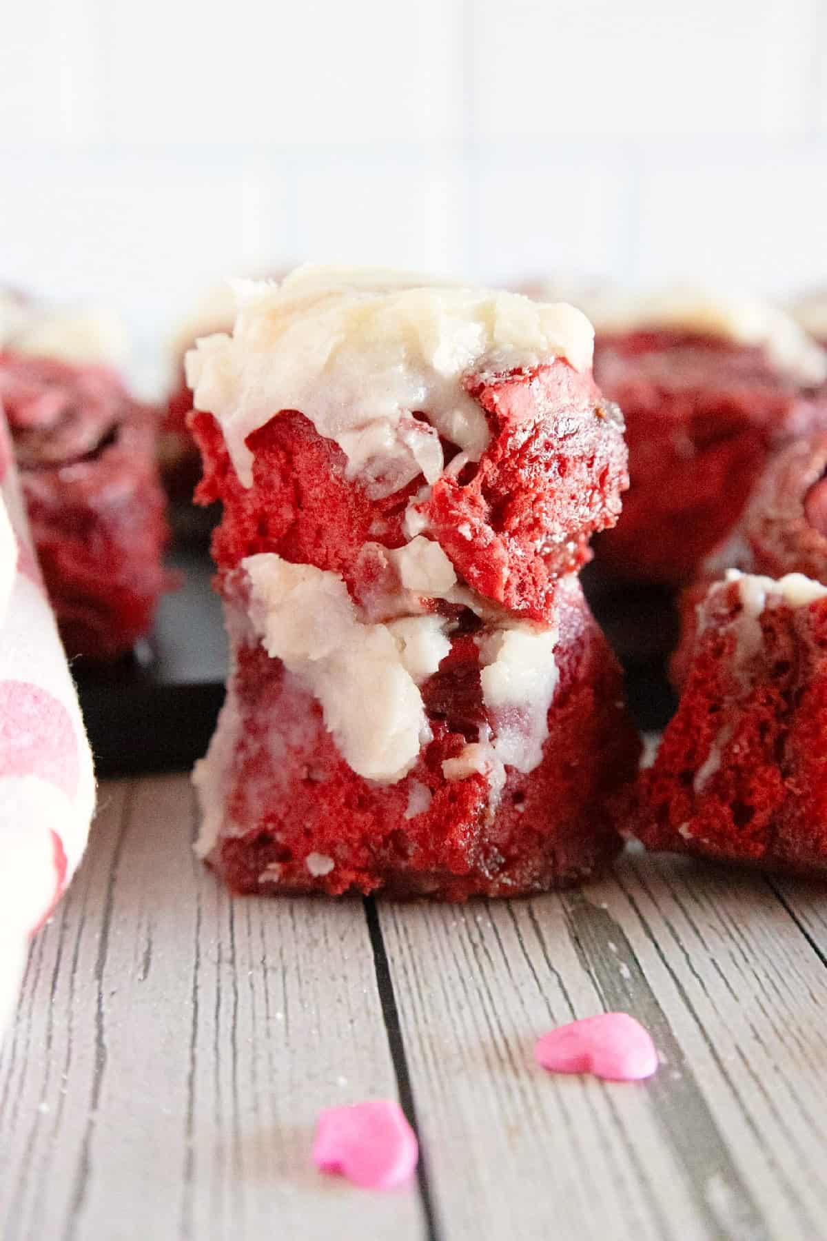 Frosted red velvet cinnamon roll served with heart sprinkles on a rustic wood surface.