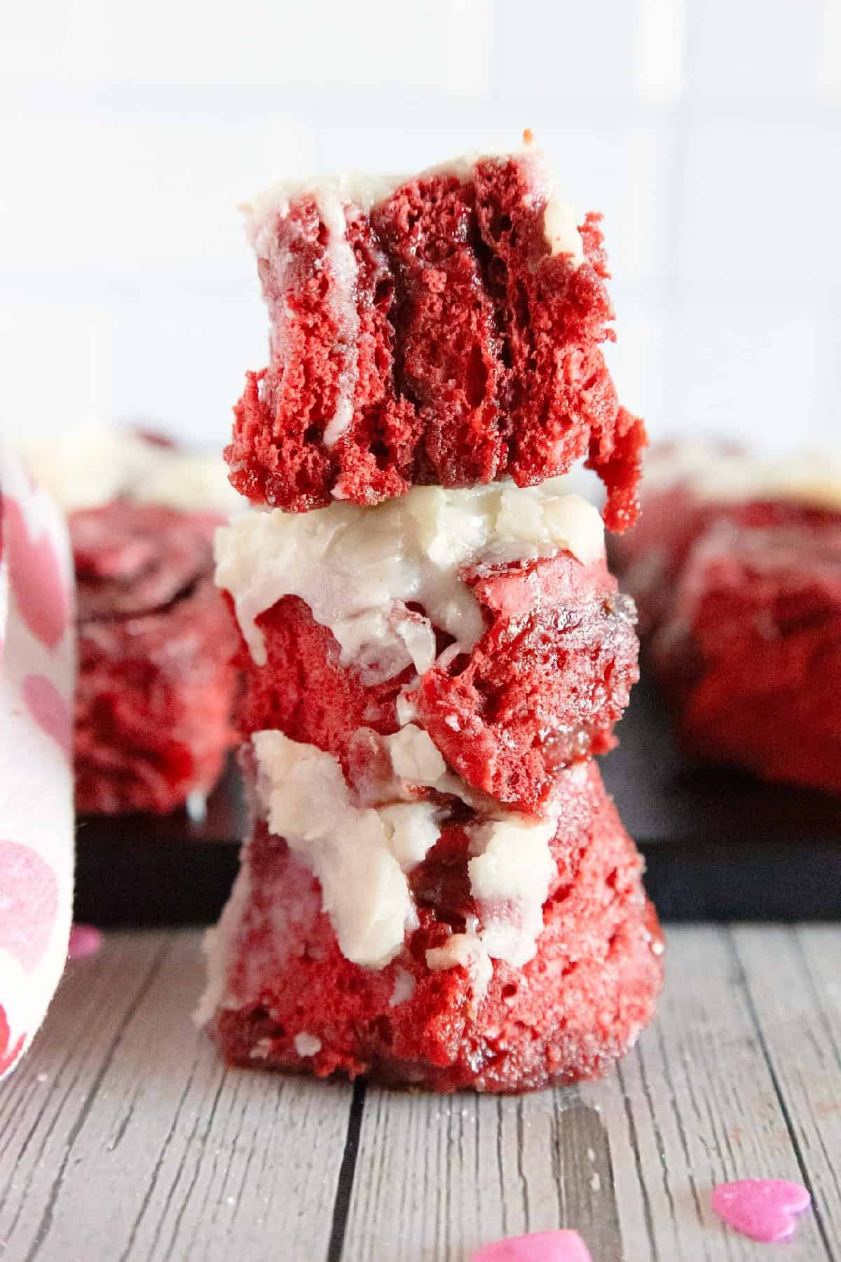 Stacked red velvet cinnamon rolls with cream cheese frosting and heart sprinkles.