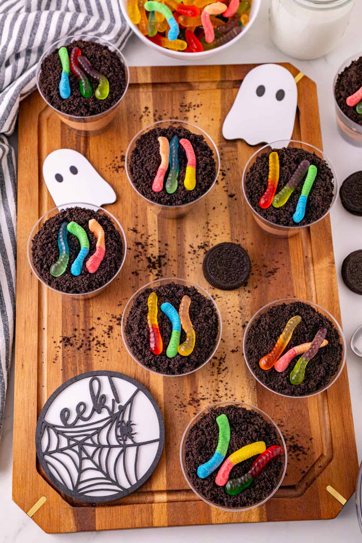 Dirt and worms pudding cups topped with Oreo crumbs and colorful gummy worms on a Halloween serving board.