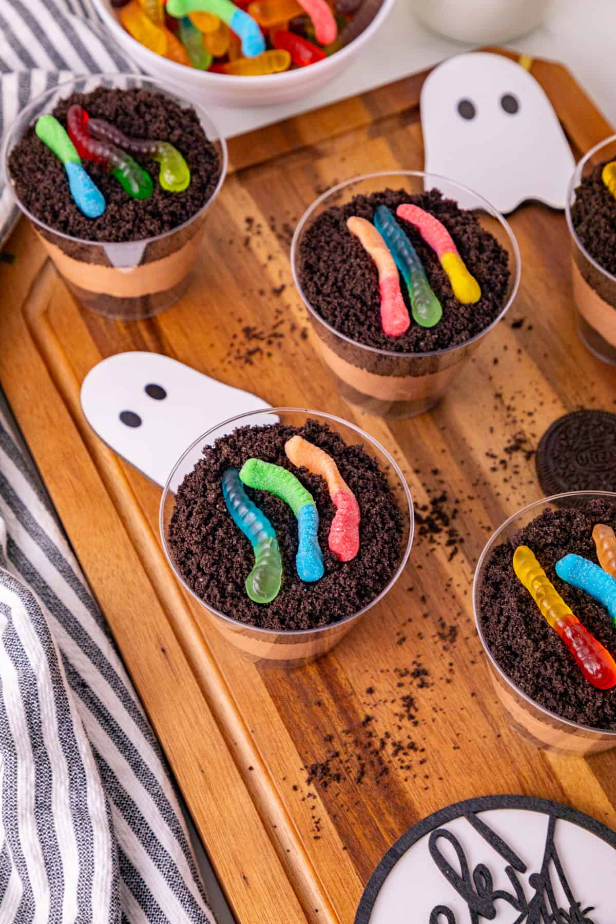 Dirt and worms pudding cups topped with colorful gummy worms and Oreo crumbs on a wooden serving board.